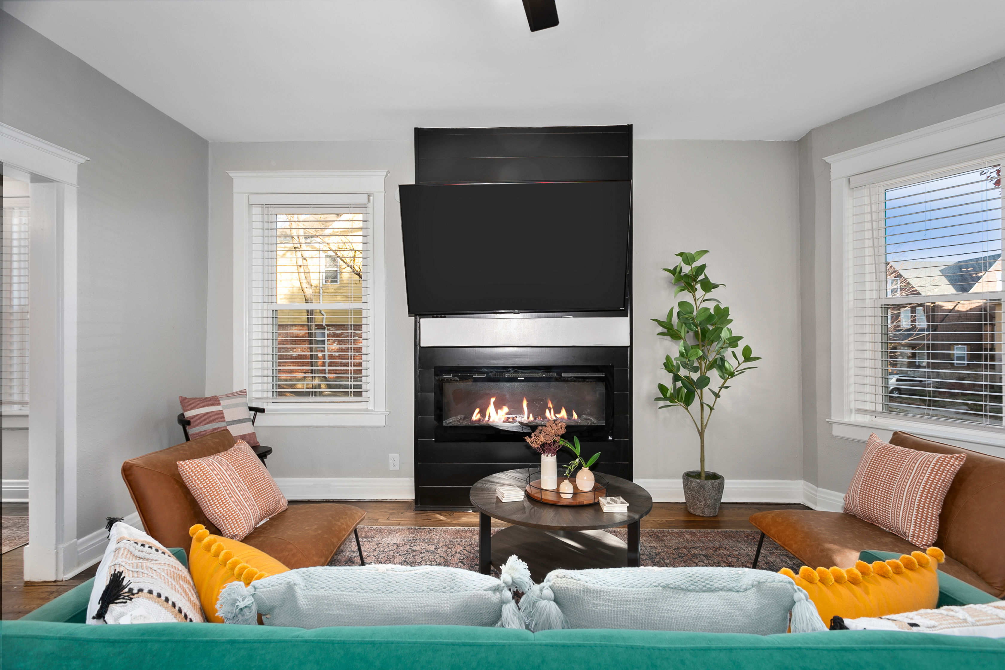 Cozy modern living room with wall-mounted TV above a black gas fireplace, teal sofa with colorful pillows, leather accent chairs, round coffee table, potted plant, hardwood floors and twin windows with blinds