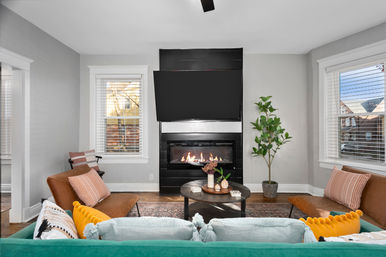 Cozy modern living room with wall-mounted TV above a black gas fireplace, teal sofa with colorful pillows, leather accent chairs, round coffee table, potted plant, hardwood floors and twin windows with blinds