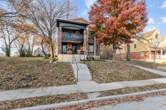 Two-story brick house with covered front balcony and central concrete steps from the sidewalk, set on a grassy slope with a red autumn tree and neighboring homes on a tree-lined residential street.