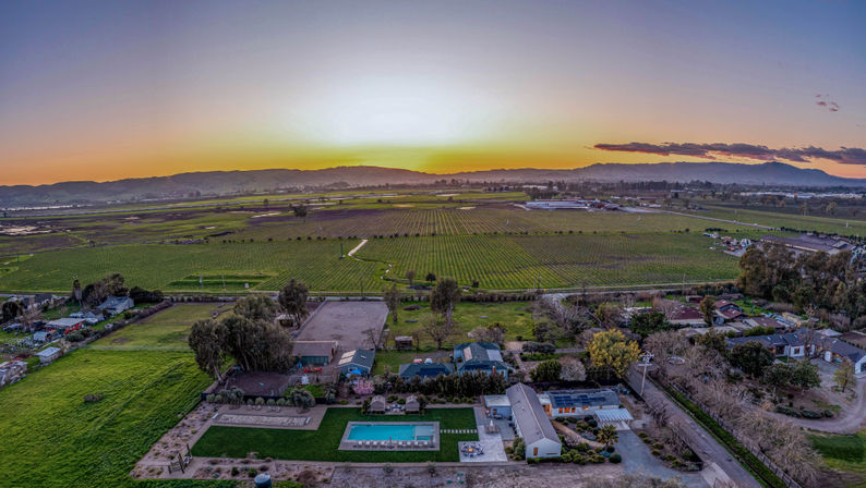 Aerial sunset view of a rural valley with neat vineyard rows and farmland stretching to rolling hills, homes and a bright backyard swimming pool in the foreground