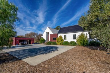 Sunlit modern farmhouse-style home with white siding and black solar panels, a red detached garage, gravel driveway and trimmed shrubs beneath a vivid blue sky.
