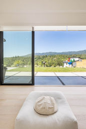 Bright modern living room with low white cushion and textured hat facing floor-to-ceiling sliding glass doors opening to a patio and lawn, colorful rooster garden sculpture, and tree‑covered rolling hills under a clear blue sky.