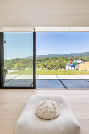 Bright modern living room with low white cushion and textured hat facing floor-to-ceiling sliding glass doors opening to a patio and lawn, colorful rooster garden sculpture, and tree‑covered rolling hills under a clear blue sky.