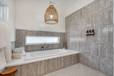 Spa-style modern bathroom with built-in white soaking tub set in glossy vertical gray subway tile, narrow horizontal window, mosaic tile floor, woven rattan pendant light, stacked white towels and wall-mounted soap dispensers.