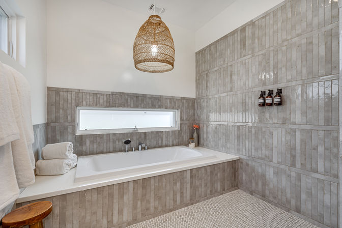 Spa-style modern bathroom with built-in white soaking tub set in glossy vertical gray subway tile, narrow horizontal window, mosaic tile floor, woven rattan pendant light, stacked white towels and wall-mounted soap dispensers.