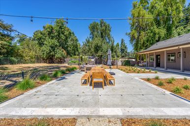 Sunlit backyard concrete patio with a long wooden dining table and benches, string lights overhead, lounge seating and umbrella, landscaped yard and mature trees near a covered porch