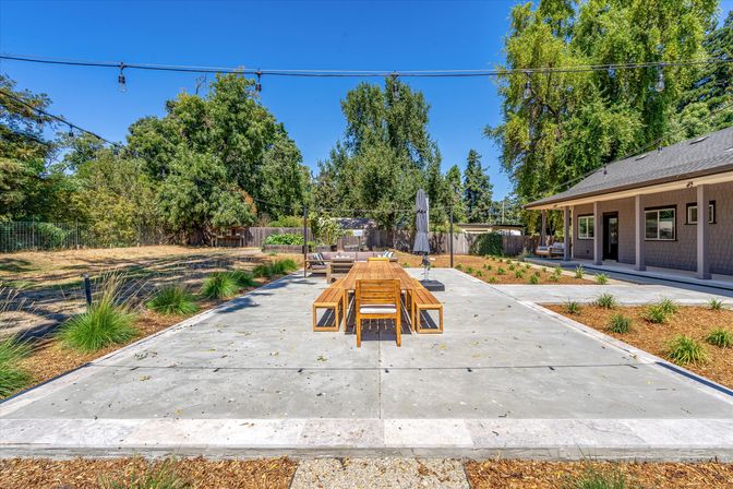 Sunlit backyard concrete patio with a long wooden dining table and benches, string lights overhead, lounge seating and umbrella, landscaped yard and mature trees near a covered porch