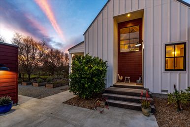 Cozy modern farmhouse entrance at sunset — white board-and-batten exterior with a deep-red recessed door, concrete steps flanked by potted plants and a lemon tree, warm interior light glowing through windows and raised garden beds under a colorful sky.