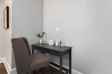 Cozy home office corner with gray textured walls, a dark wood two-drawer desk topped with a small plant, ceramic vase, tissue box and bottle, and a brown upholstered swivel chair on hardwood floors.
