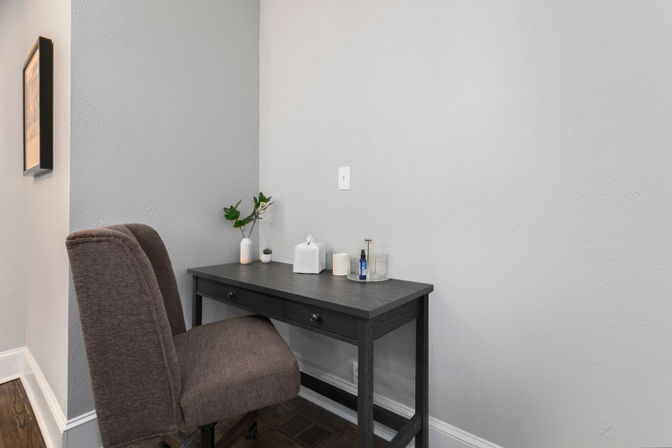 Cozy home office corner with gray textured walls, a dark wood two-drawer desk topped with a small plant, ceramic vase, tissue box and bottle, and a brown upholstered swivel chair on hardwood floors.