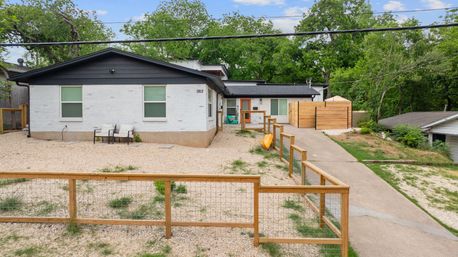 Front exterior of a modern white brick ranch house with black trim and an orange front door, gravel yard with low wooden fence, concrete driveway, and trees in the background.