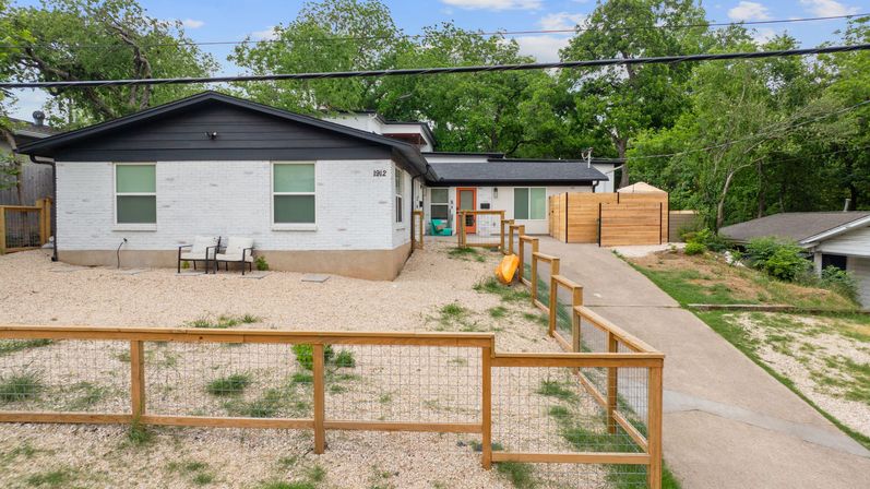 Front exterior of a modern white brick ranch house with black trim and an orange front door, gravel yard with low wooden fence, concrete driveway, and trees in the background.