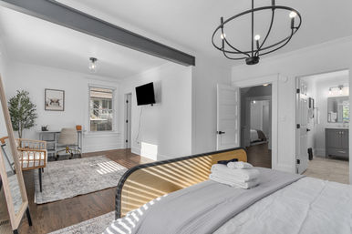 Sunlit modern master bedroom with hardwood floors, metal chandelier, bed with folded towels, wall-mounted TV and a desk nook leading to an ensuite bathroom