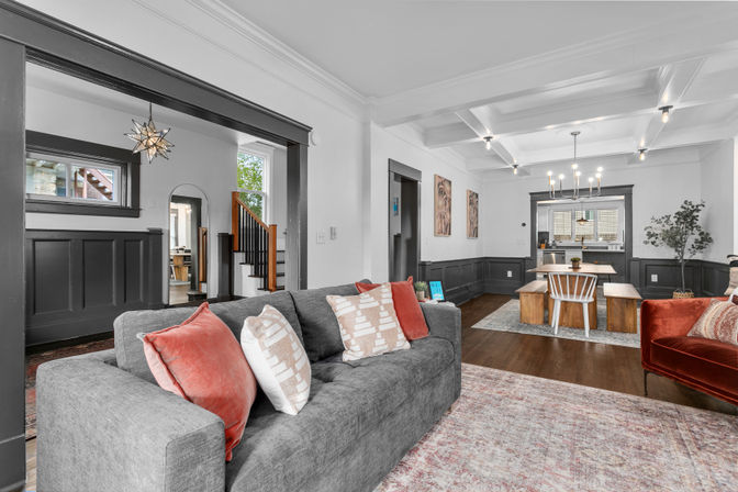 Open-concept living and dining room with a gray sofa and coral pillows, dark wainscoting and trim, coffered ceiling, wood floors, wooden dining table with benches under a chandelier, star pendant light near a staircase.
