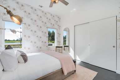 Sunlit modern bedroom with dandelion-pattern wallpaper, white bedding and pale pink throw on a wooden bedframe, brass wall lamps, arched full-length mirror, sliding closet doors, and windows overlooking a backyard pool and green landscape