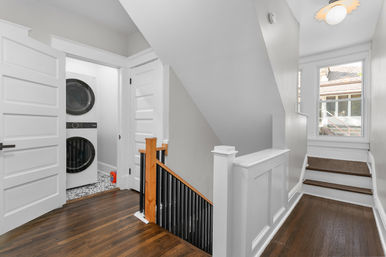 Sunlit upstairs home landing with hardwood floors, white trim, staircase with wood post and black spindles, open laundry closet showing stacked washer and dryer, and a window overlooking the neighborhood.