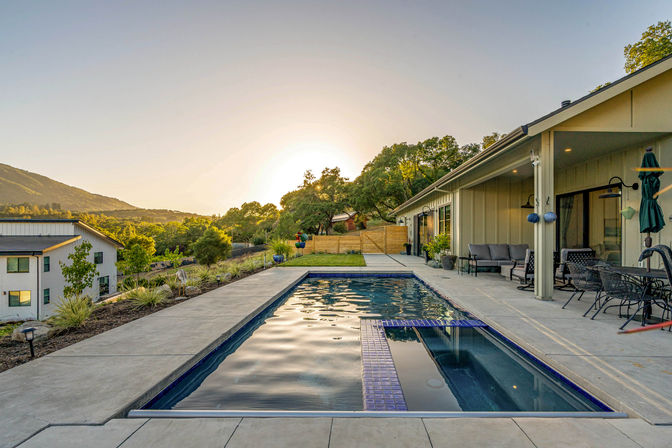 Rectangular lap pool beside a modern ranch-style house patio with outdoor seating, potted plants and rolling hills at sunset