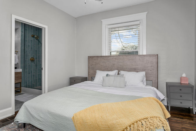 Cozy modern bedroom interior with light gray walls, wooden headboard, white bedding, pale green quilt and yellow throw, gray nightstands, hardwood floors, window blinds, and an ensuite teal-tiled shower visible through the doorway.