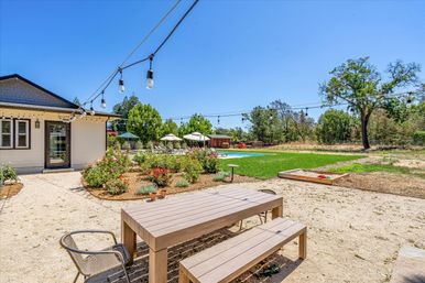 Sunny residential backyard entertaining space with wooden picnic table and benches, hanging string lights, rose garden, lawn, pool with umbrellas and lounge chairs, sandbox, and trees under a clear blue sky.
