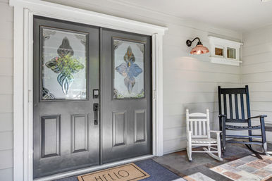 Welcoming covered front porch with double black entry doors featuring decorative stained-glass panels, a "HELLO" doormat, black adult and white child rocking chairs, and a copper wall sconce on gray siding.