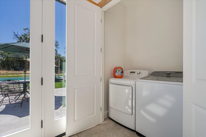 Sunny laundry nook with white washer and dryer and orange detergent bottle on top, open glass French doors leading to patio dining set, umbrella, pool and trees