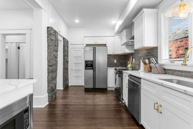 Bright modern white galley kitchen with stainless steel appliances, marble-look countertops, gray subway tile backsplash, dark hardwood floors, exposed stone column accents and a sunny window with blinds.