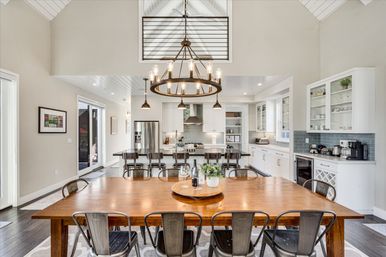 Open-concept modern farmhouse kitchen and dining area with vaulted ceiling and round metal chandelier, large wooden dining table with metal chairs, breakfast island with barstools, white cabinetry and stainless appliances