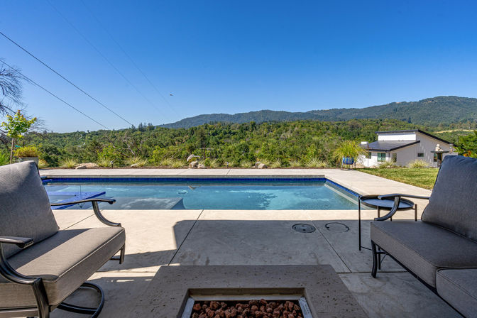 Backyard pool and patio with cushioned lounge chairs and a fire pit, overlooking green hills and distant mountains under a clear blue sky