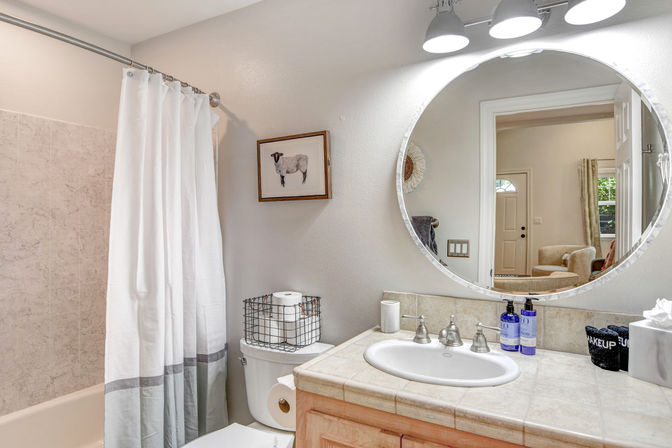 Bright neutral bathroom with round mirror reflecting a hallway, tiled vanity with sink and chrome faucet, blue soap bottles on the counter, white shower curtain over tub, toilet topped with a wire basket of toilet paper and a framed sheep print on the wall.
