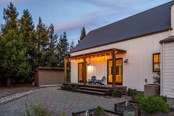 Twilight modern farmhouse exterior with white board-and-batten siding, cozy covered wooden porch strung with warm globe lights, two Adirondack chairs, gravel yard, garden beds, storage shed and tall evergreen trees.