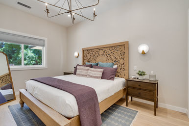 Cozy contemporary bedroom interior with ornate carved wooden headboard, king-size bed in white linens with purple throw and decorative pillows, matching wood nightstands with sconces, large window showing green trees, full-length mirror and light hardwood floors.
