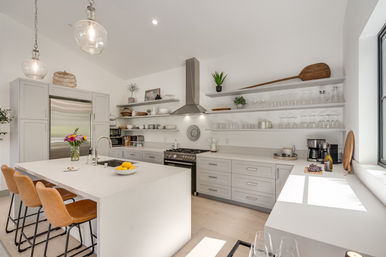 Sunlit modern white kitchen with waterfall island and three tan leather bar stools, stainless steel refrigerator and range, open floating shelves holding dishes and glassware, pendant globe lights and wood accent decor.