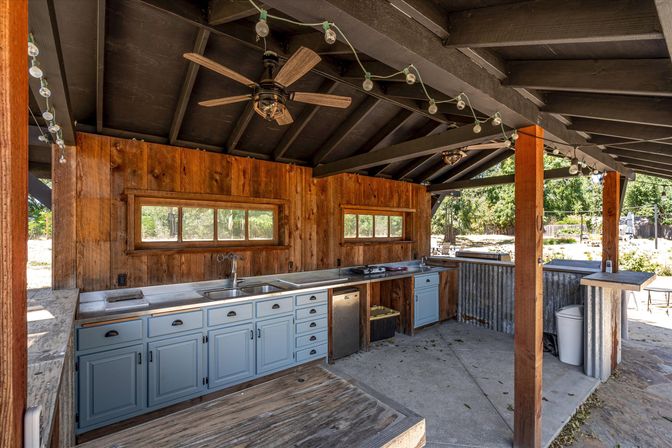 Rustic backyard outdoor kitchen on covered patio with black beamed ceiling, ceiling fans and string lights, stainless sink and stove, pale blue cabinets and corrugated metal bar