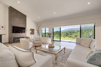 Sunlit modern living room with neutral sofas and armchairs around a glass-top coffee table, wall-mounted TV above a sleek fireplace, and floor-to-ceiling sliding glass doors revealing a panoramic green mountain view and patio.