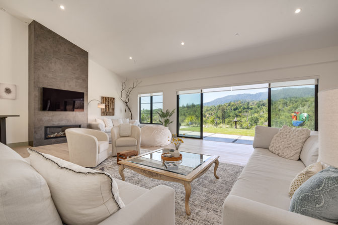 Sunlit modern living room with neutral sofas and armchairs around a glass-top coffee table, wall-mounted TV above a sleek fireplace, and floor-to-ceiling sliding glass doors revealing a panoramic green mountain view and patio.