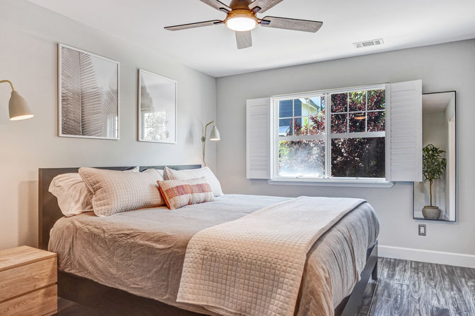 Cozy sunlit modern master bedroom with king bed, neutral quilted bedding and accent pillow, wooden nightstand, ceiling fan, plantation shutters framing a leafy view, framed wall art and potted plant on gray wood floors.