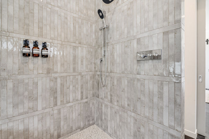 Spa-like walk-in shower with vertical gray subway-style tiles, chrome rainfall and handheld showerhead, wall-mounted shampoo dispensers, and white hexagon floor tiles.