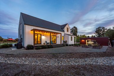 Modern white farmhouse with dark metal roof and covered wooden porch lit by warm string lights at dusk, gravel courtyard with raised garden beds and an open detached garage in a peaceful rural setting.