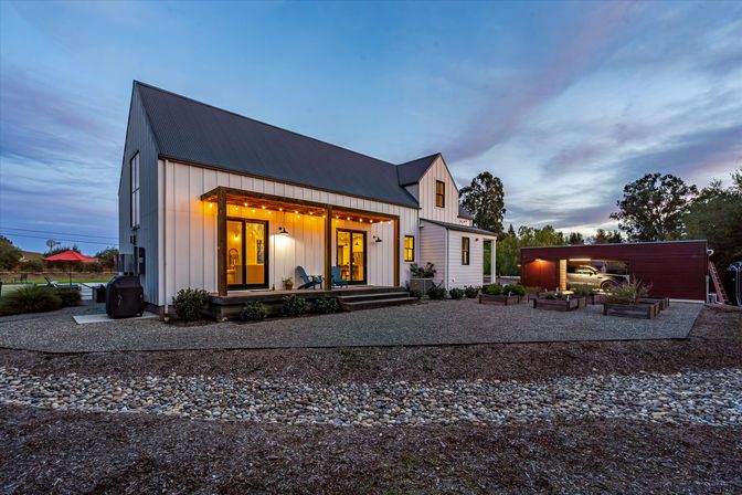 Modern white farmhouse with dark metal roof and covered wooden porch lit by warm string lights at dusk, gravel courtyard with raised garden beds and an open detached garage in a peaceful rural setting.
