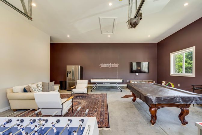 Spacious home game room with maroon accent wall, covered wooden pool table, foosball table in foreground, air hockey table, comfy sofa and chairs on a patterned rug, stainless refrigerator, wall-mounted TV, concrete floor, garage door opener overhead and window showing backyard greenery