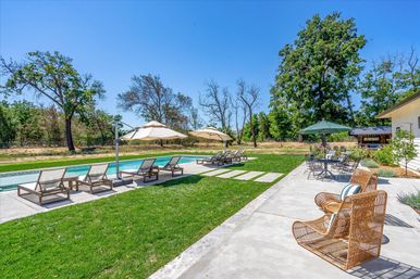 Sunny residential backyard with a rectangular swimming pool, rows of lounge chairs under beige umbrellas on a concrete deck, wicker patio chairs and a dining table under a green umbrella, lush lawn and mature trees beneath a clear blue sky.