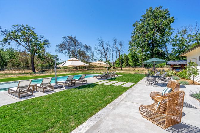 Sunny residential backyard with a rectangular swimming pool, rows of lounge chairs under beige umbrellas on a concrete deck, wicker patio chairs and a dining table under a green umbrella, lush lawn and mature trees beneath a clear blue sky.