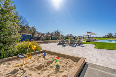 Sun-drenched backyard pool area with a wooden sandbox and yellow digger toy in the foreground, paved patio with lounge chairs and umbrellas, manicured lawn and bright blue sky.