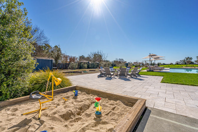 Sun-drenched backyard pool area with a wooden sandbox and yellow digger toy in the foreground, paved patio with lounge chairs and umbrellas, manicured lawn and bright blue sky.