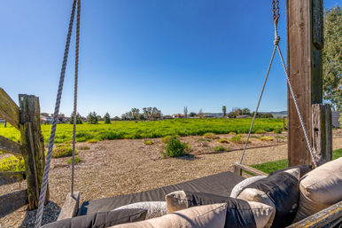 Inviting outdoor swing bed with cushions hung from wooden posts and ropes, overlooking a gravel path, bright green field, distant houses and rolling hills under a clear blue sky