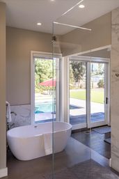 Modern bathroom with a freestanding white soaking tub, glass shower partition and marble accents, sliding glass doors opening to a sunny backyard with a pool and red patio umbrella.
