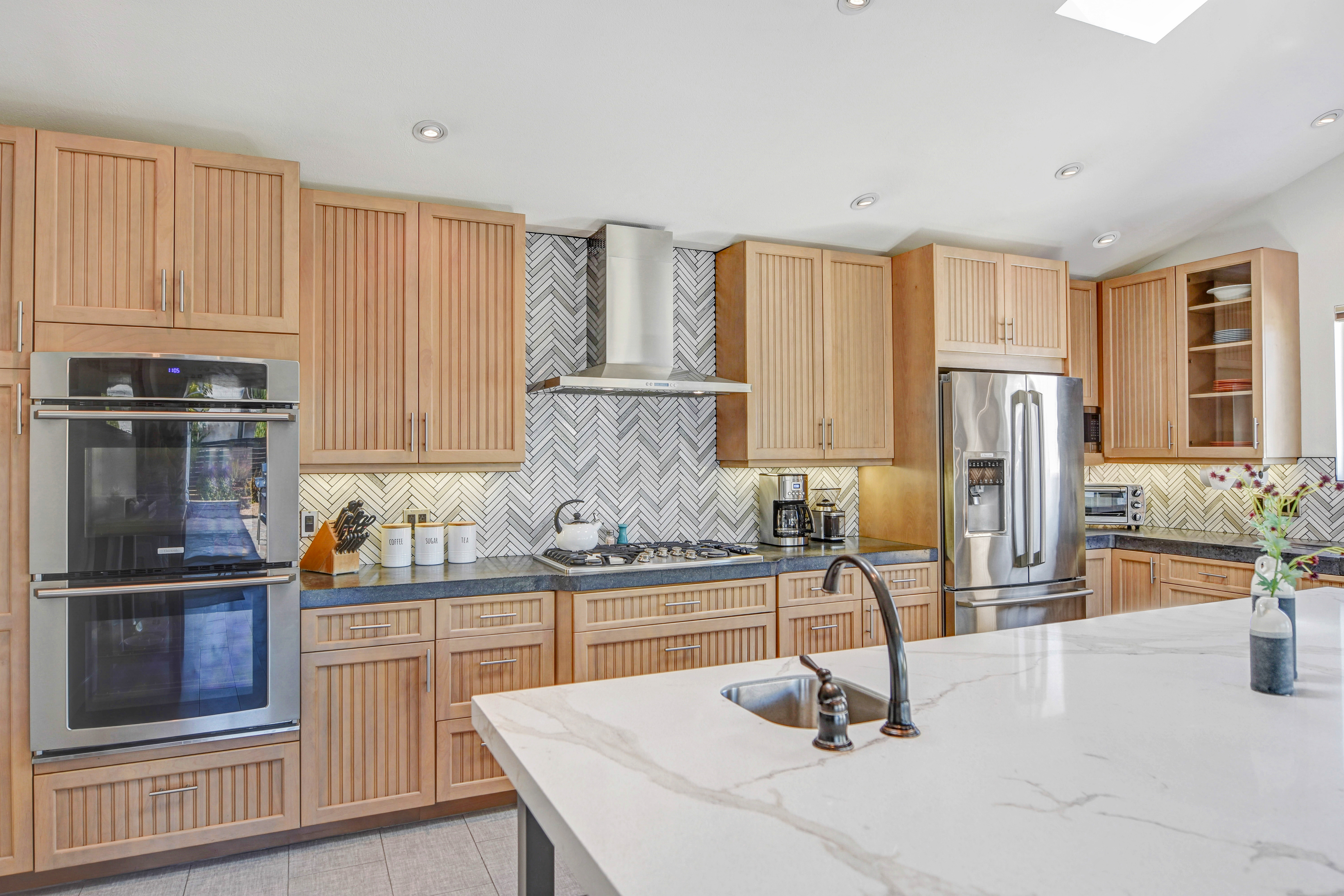Bright modern kitchen with light wood beadboard cabinets, stainless steel double oven and refrigerator, herringbone tile backsplash, gas cooktop under a stainless hood, and a white marble island with built-in sink and dark faucet.