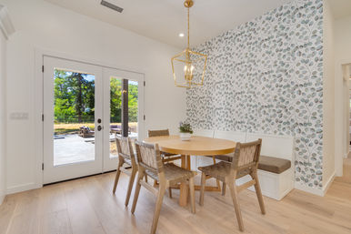Sunlit dining nook with round light-wood table, woven chairs and built-in banquette, leaf-pattern wallpaper and French doors to a patio.