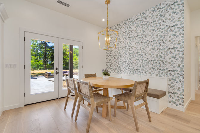 Sunlit dining nook with round light-wood table, woven chairs and built-in banquette, leaf-pattern wallpaper and French doors to a patio.