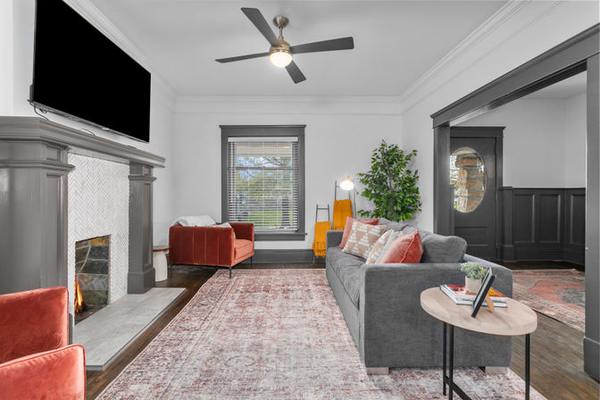 Cozy craftsman-style living room with gray trim, lit fireplace and wall-mounted TV, ceiling fan, gray sofa with patterned pillows, rust velvet accent chairs, large patterned area rug, window and oval-paneled entry door.